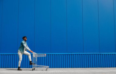 Siyah afro adam alışveriş arabasıyla alışverişe gidiyor, adam eğleniyor ve zıplıyor, alışveriş merkezinin yakınında alışverişler için koşuyor, mavi bir binanın arka planında genç bir zenci öğrenci var.