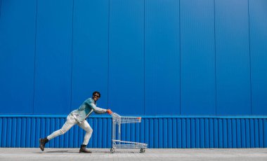 Siyah afro adam alışveriş arabasıyla alışveriş yapıyor. Eğleniyor ve zıplıyor. Alışveriş merkezinin yakınında alışveriş yapıyor.