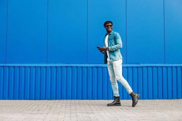 african guy walks with a phone around the city, against the background of a blue building, makes a purchase online through the phone, a full-length portrait