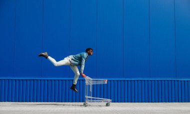 Siyah afro adam alışveriş arabasıyla alışveriş yapıyor. Eğleniyor ve zıplıyor. Alışveriş merkezinin yakınında alışveriş yapıyor.