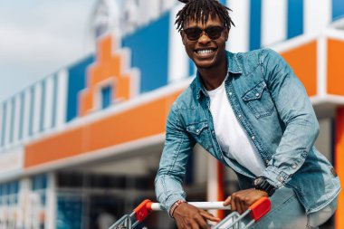 Afro-Amerikan siyah adam alışveriş merkezinin önünde alışveriş arabasıyla alışveriş yapıyor başarılı bir alışveriş merkezi, güneş gözlüğü takıyor.