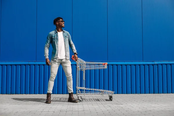 Siyah afro adam alışveriş arabasıyla alışveriş yapıyor, adam poz veriyor, moda moda, alışveriş merkezinin yakınında alışveriş yapıyor, mavi bir binanın arka planında genç bir zenci öğrenci var.
