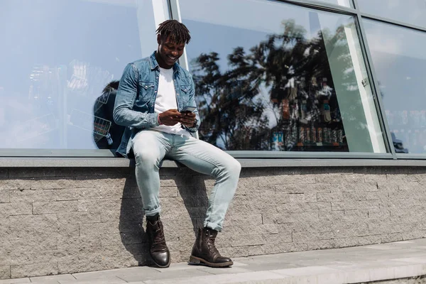 black man African American with a phone in his hands, typing an SMS message, sitting against an educational institution, in the city center, against a glass building, cheerful African