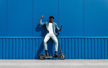 Elektrikli scooterlı siyah Afro-Amerikan bir adam, elinde bir telefon tutarken bir başarı jesti gösteriyor, şehirdeki mavi bir duvarın arka planında, 5 gramlık internet hızında