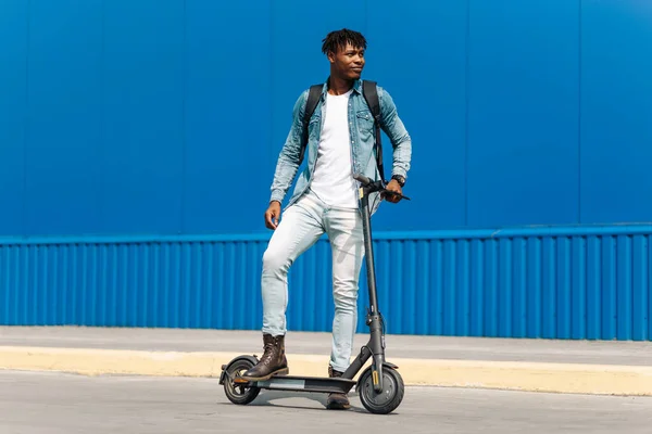 骑电动车的年轻的非洲裔美国人 骑在路上 在城市蓝色建筑的背景下 奔向学校或工作 摆出与摩托车并排的架势 — 图库照片