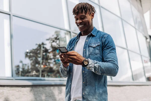 black man African American with a phone in his hands, typing an SMS message, walking in the city center, against a glass building, cheerful African with a phone in his hands