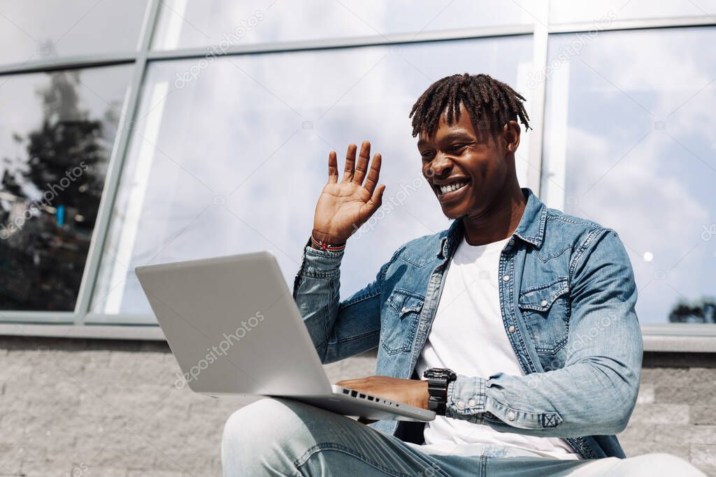 black african american man typing on a laptop on the street in front of a glass building sitting, talking skype, high-speed mobile internet, 5g, 4g, waves his hand hello to the interlocutor