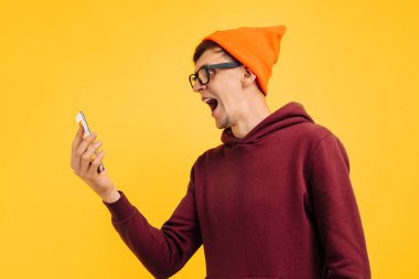 Gözlüklü, kırmızı kazaklı, turuncu şapkalı kızgın genç adam telefonda yüksek sesle konuşuyor.