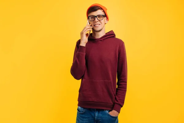 オレンジの帽子の眼鏡と赤いセーターのハンサムな若い男が電話で話し 黄色の背景 秋のコンセプトに立っている間に笑う — ストック写真
