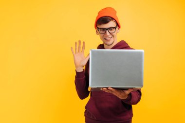 Genç ve yakışıklı bir adam internetten iletişim kuruyor, bir laptop, bir video çağrısı, turuncu şapkalı ve kırmızı kazaklı, sarı arka planda, yüksek hızlı internet, 5 G
