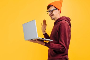 Genç ve yakışıklı bir adam internetten iletişim kuruyor, bir laptop, bir video çağrısı, turuncu şapkalı ve kırmızı kazaklı, sarı arka planda, yüksek hızlı internet, 5 G