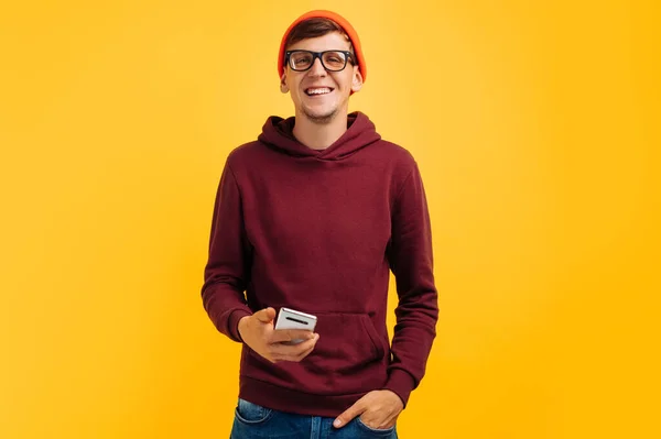 眼鏡をかけたオレンジの帽子のハンサムな若い男と赤いセーターが電話にメッセージを入力して手に電話を持ち黄色の背景に立っている秋のコンセプト5G — ストック写真