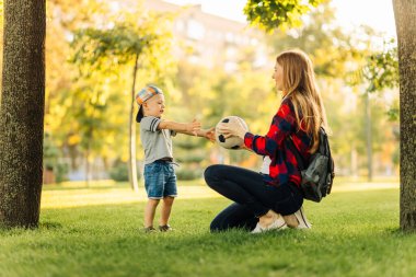 Genç mutlu anne ve küçük oğlu parkta futbol oynuyorlar, anne oğluna futbol topu veriyor, eğleniyor ve güneşli bir yaz gününde topla oynuyor.