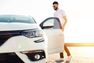 Şehir merkezinde bir arabanın yanında duran güneş gözlüklü şık, genç ve yakışıklı bir adam. Şehir caddesinde, modern beyaz bir arabanın yanında duran yakışıklı adam.