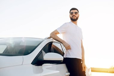 Yakışıklı genç bir adam, güneş gözlüklü ve beyaz gömlekli bir işadamı, spor arabasının yanında duruyor, arabasının yanında, dışarıda.