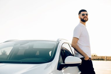 Yakışıklı genç bir adam, güneş gözlüklü ve beyaz gömlekli bir işadamı, spor arabasının yanında duruyor, arabasının yanında, dışarıda.