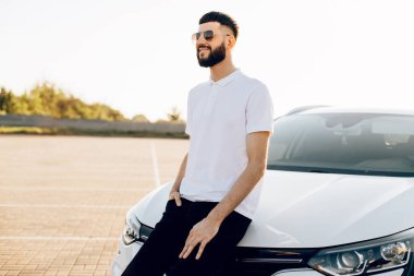 Güneş gözlüklü, sakallı, lüks arabalı, arabanın yanında mutlu bir adamın portresi. Seyahat, ulaşım, tatil.