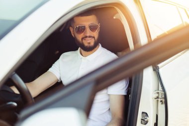 Mutlu sahip, yakışıklı sakallı genç adam, beyaz tişörtlü, yeni arabasında rahatlamış, pencereden dışarı bakıyor neşeyle gülümsüyor.
