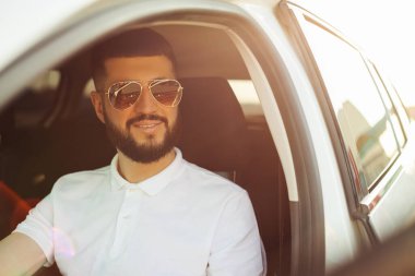 Mutlu sahip, yakışıklı sakallı genç adam, beyaz tişörtlü, yeni arabasında rahatlamış, pencereden dışarı bakıyor neşeyle gülümsüyor.