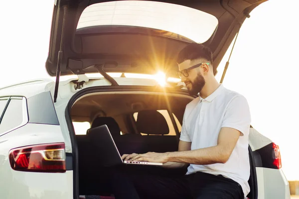 Young man in sunglasses enjoying a trip in his car, sitting in the trunk of a car with a laptop, Travel concept, summer vacation, vacation