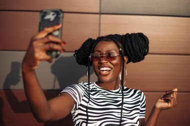 Güzel bir Afro-Amerikan kadının portresi, güneş gözlüğü takıyor, gülümseyen bir kadın cep telefonuyla selfie çekiyor, Selfie konsepti..