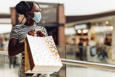 Alışveriş merkezinde koruyucu maske takan Afrikalı genç bir kadın alışveriş torbaları, alışveriş konsepti, karantina, COVID-19