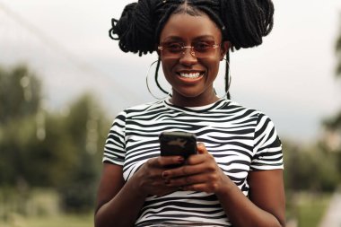 Afro saç stili olan tatmin olmuş hippi siyahi kadın cep telefonunda mesaj yazıyor açık havada sohbet etmekten hoşlanıyor, teknoloji kavramı.