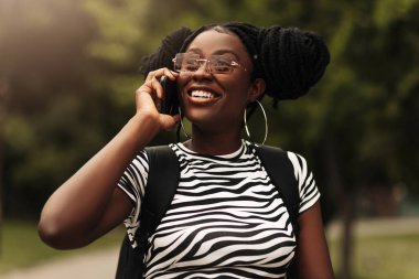 Gülümseyen Afrikalı Amerikalı kadın, güneş gözlüğü takıyor, sokakta yürürken cep telefonuyla konuşuyor.