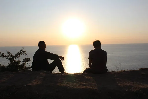 silhouette of two men sitting on the beach looking at the sunset / photo two silhouettes of men. they are sitting on the beach. watching the sunset. there is a Sunny path on the sea.