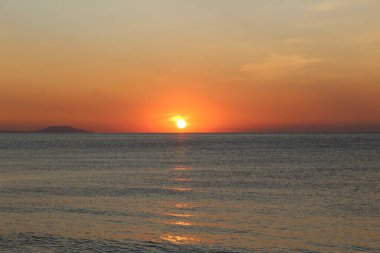 Denizin üzerinde gün batımı ve su üzerinde güneşli bir yol. Gün batımı turuncu fotoğraf çemberi. Gökyüzünde kırmızı bir parıltı var. Deniz sakindir. Su mavi..