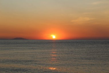 Denizin üzerinde gün batımı ve su üzerinde güneşli bir yol. Gün batımı turuncu fotoğraf çemberi. Gökyüzünde kırmızı bir parıltı var. Deniz sakindir. Su mavi..