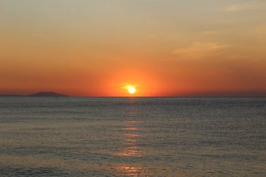 Denizin üzerinde gün batımı ve su üzerinde güneşli bir yol. Gün batımı turuncu fotoğraf çemberi. Gökyüzünde kırmızı bir parıltı var. Deniz sakindir. Su mavi..