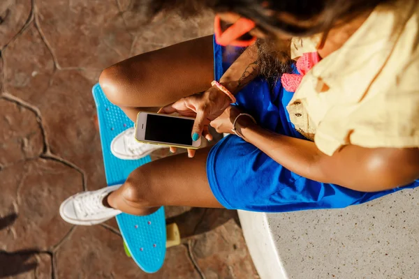 Belle adolescente noire en jupe bleue s'asseoir avec son smartpho — Photo