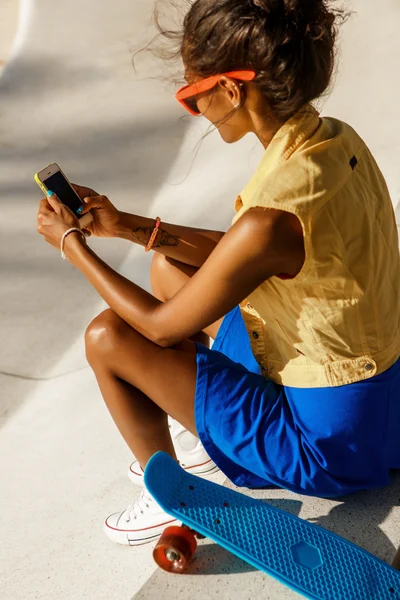 Schöne schwarze Teenager-Mädchen in blauem Rock sitzen mit ihrem Smartphone — Stockfoto