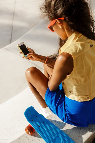 Belle adolescente noire en jupe bleue s'asseoir avec son smartpho — Photo
