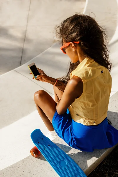 Schöne schwarze Teenager-Mädchen in blauem Rock sitzen mit ihrem Smartphone — Stockfoto