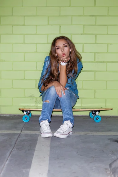 Schöne langhaarige Mädchen mit einem hölzernen Skateboard in der Nähe eines grünen — Stockfoto