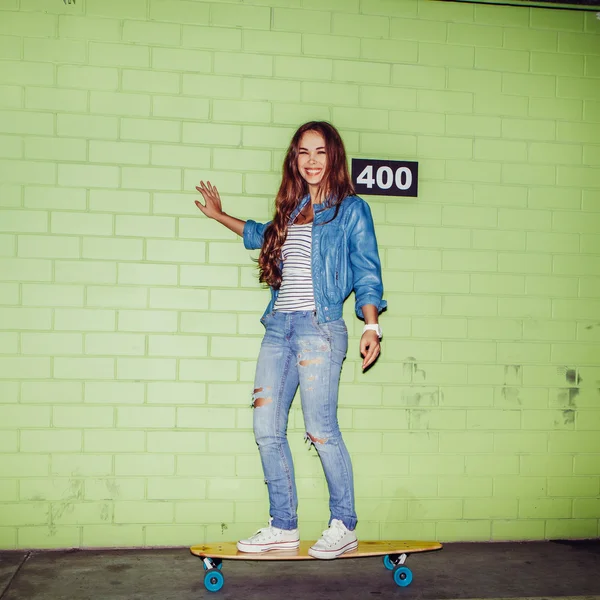 Schöne langhaarige Mädchen mit einem hölzernen Skateboard in der Nähe eines grünen — Stockfoto