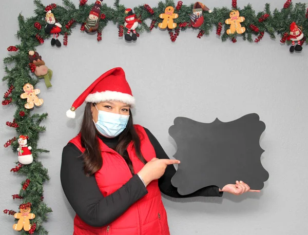 latin woman with protection mask, santa claus hat and blackboard for message.Christmas decoration, new normal covid-19
