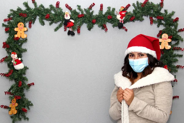 latin woman with protection mask, sweater and christmas hat on gray background with christmas decoration, new normal