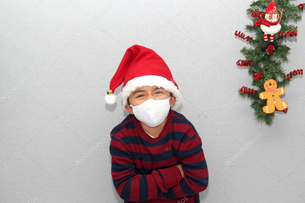 7-year-old latin boy with protection mask, sweater and Christmas hat on gray background with Christmas decoration, new normal covid-19