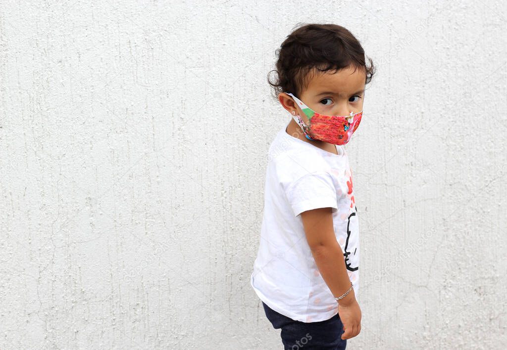 2-year-old Latin girl with protective mask on white wall background, new normal covid-19