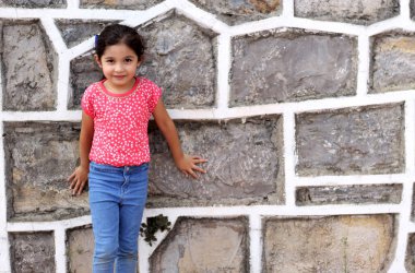 6 yaşında, latin bir kız çocuğu. Kauvid-19 koruma maskesi takmış, taş duvar arkası, yeni normal.