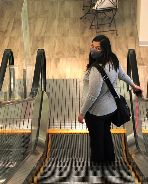 Latino woman with protection mask on escalators in shopping mall, new normal