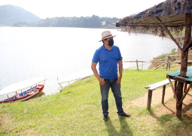 Şapkalı ve koruyucu maskeli Latin turist, sahil ve göl manzaralı.