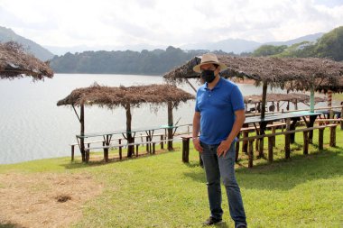 Şapkalı ve koruyucu maskeli Latin turist, sahil ve göl manzaralı.
