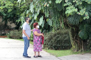Anne ve oğul, kızıl saçlı büyükanne koruyucu maskeler takan Latin yetişkin bir adamla kol kola yürüyor bitki ve bitki örtüsü.