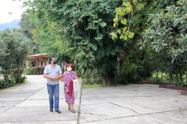 Anne ve oğul, kızıl saçlı büyükanne koruyucu maskeler takan Latin yetişkin bir adamla kol kola yürüyor bitki ve bitki örtüsü.