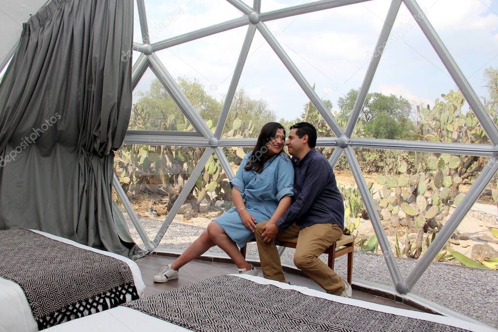 Latin couple in love inside glamping dome room with desert vegetation outside