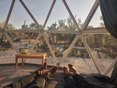 Çift yatağında uzanmış güneşin doğuşunu jeodezik çadır penceresinden izliyor.